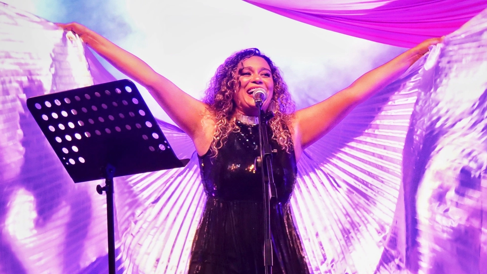 solange-cesarovna-on-stage-73018 Solange Cesarovna performing on stage, singing into a microphone with arms outstretched, holding shimmering silver wing-like fabric. She is illuminated by vibrant purple, pink, and blue stage lighting, creating a dramatic atmosphere.